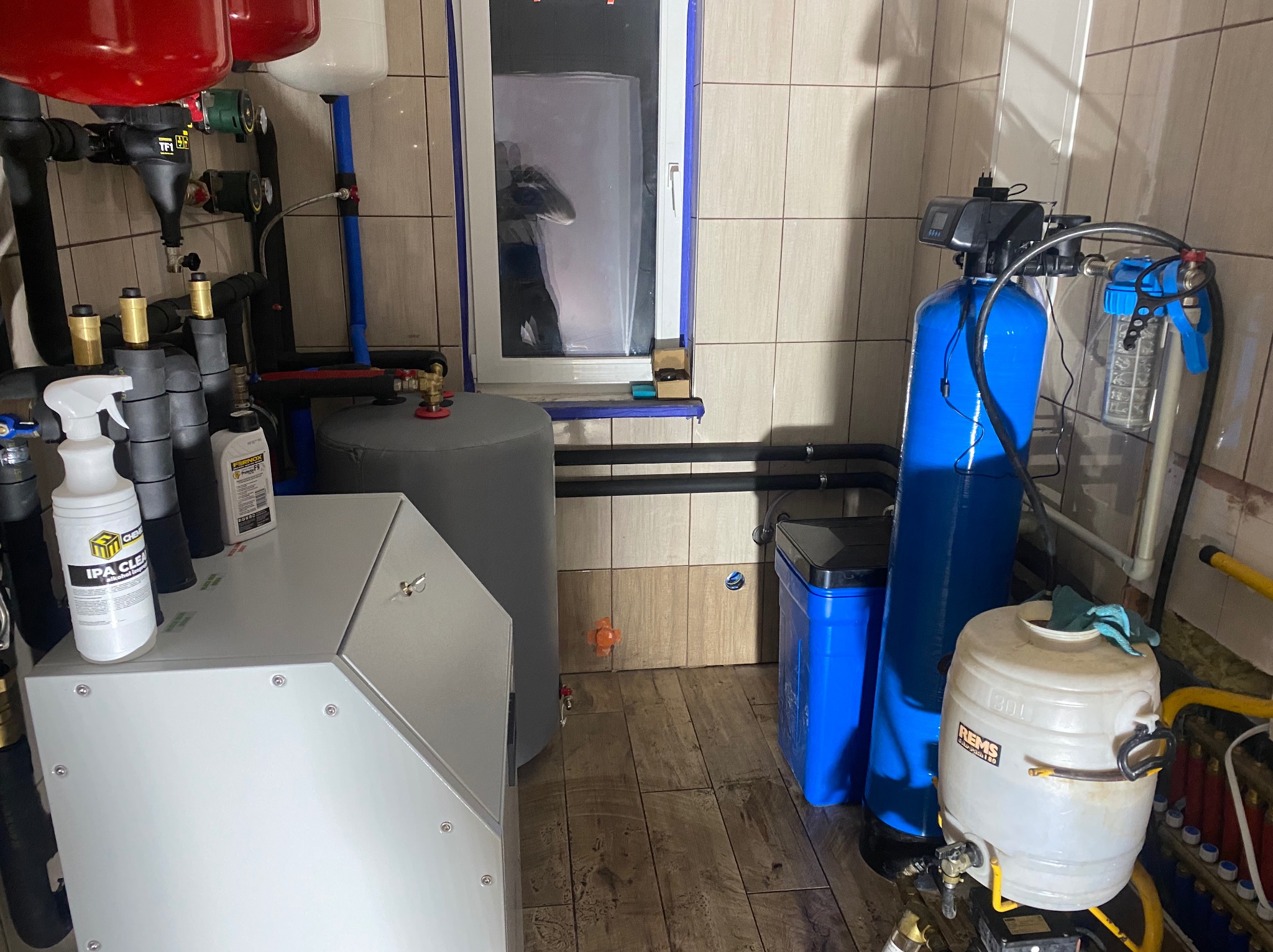 Węzeł cieplny z piecem, zbiornikiem i filtrem wody. Widoczne rury, zawory, manometry. Na piecu butelka z alkoholem izopropylowym. Tło stanowią kafelki.