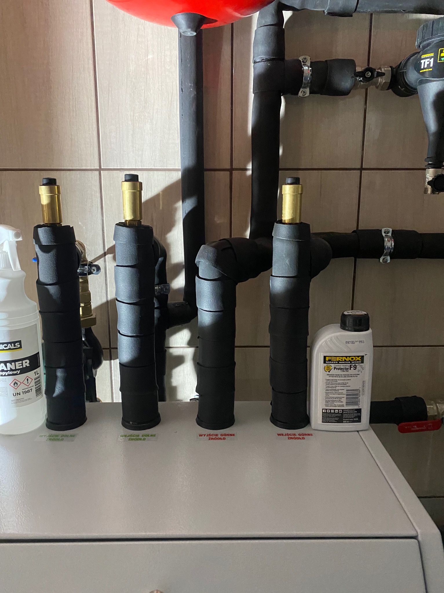 Fragment instalacji grzewczej z rurami w izolacji, z widocznymi etykietami 'wejście' i 'wyjście'. Obok stoją preparaty do czyszczenia i ochrony instalacji.