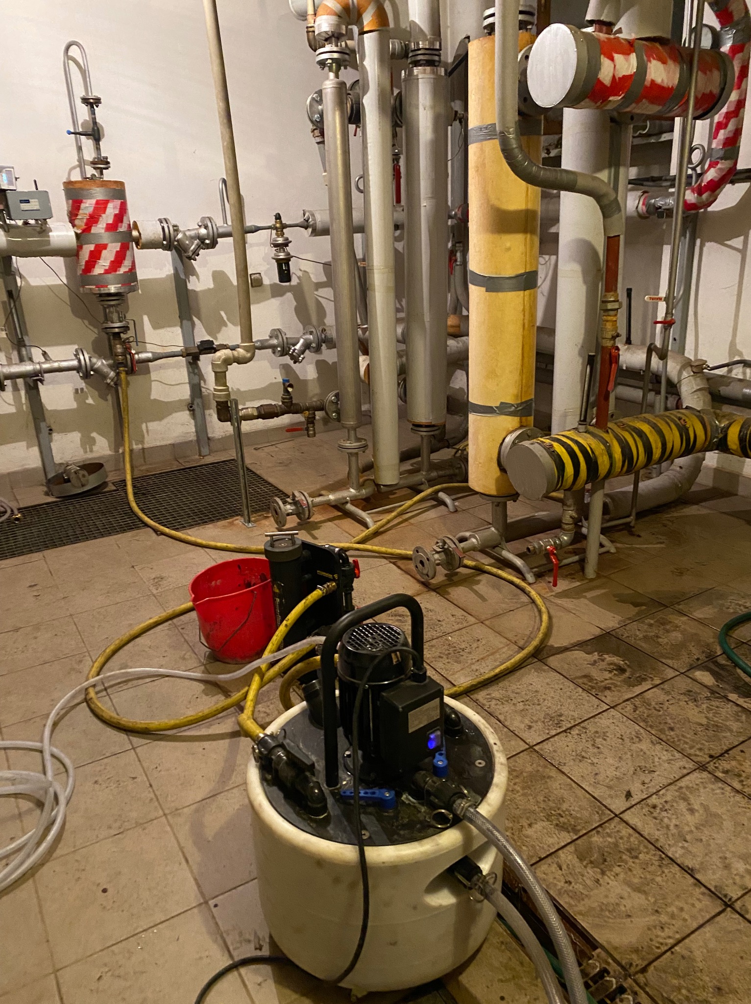 Pompa zanurzeniowa w kotłowni, podłączona do węży. W tle rury instalacji grzewczej z izolacją w kolorach czerwono-białym i żółto-czarnym.