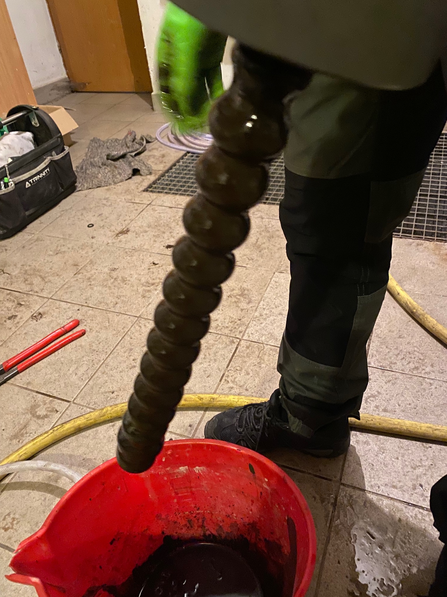 Czyszczenie rury spiralną sprężyną hydrauliczną nad czerwonym wiadrem z brudną wodą. W tle narzędzia i elementy instalacji.