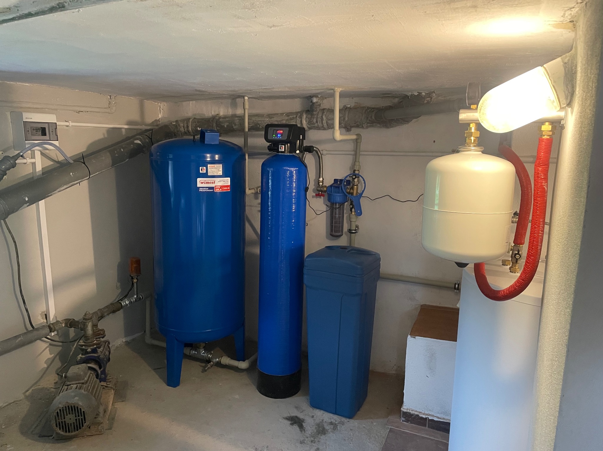Kompletna instalacja uzdatniania wody w piwnicy: dwa niebieskie zbiorniki, filtr, biały hydrofor z czerwonymi wężami, pompa i rury pod sufitem, oświetlone lampą.