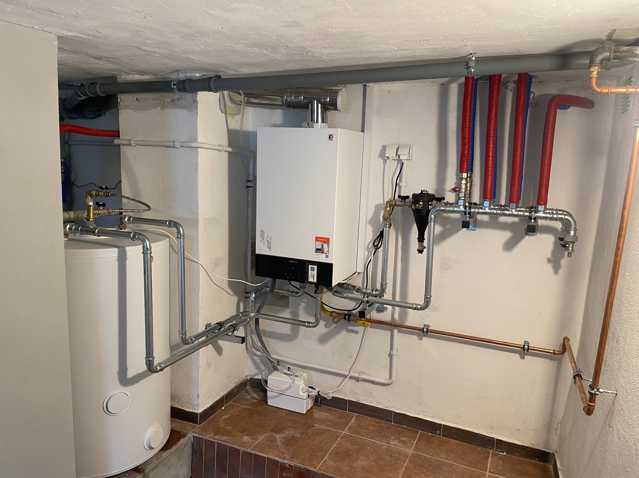 Biały piec gazowy Viessmann z szarymi rurami, czerwono-niebieskimi izolacjami i zbiornikiem na wodę w kotłowni, na tle beżowej ściany i brązowej podłogi.