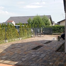 Kwasniewski Sebastian - Ułożona kostka brukowa na podjeździe, widoczne elementy montażu, ogrodzenie panelowe z zielenią, domy w tle. Częściowo ukończony projekt.