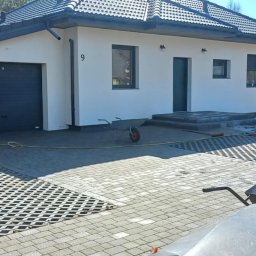 Kwasniewski Sebastian - Układanie ażurowej kostki brukowej na podjeździe domu jednorodzinnego. Widoczna ubijarka, taczki i pracownik w trakcie wykonywania prac.