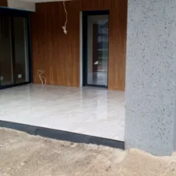 Jasne płytki na tarasie z obrzeżem, widok na kolumnę, okna i przesuwne drzwi. W tle grill i palma. Piasek wokół tarasu wskazuje na prace wykończeniowe.