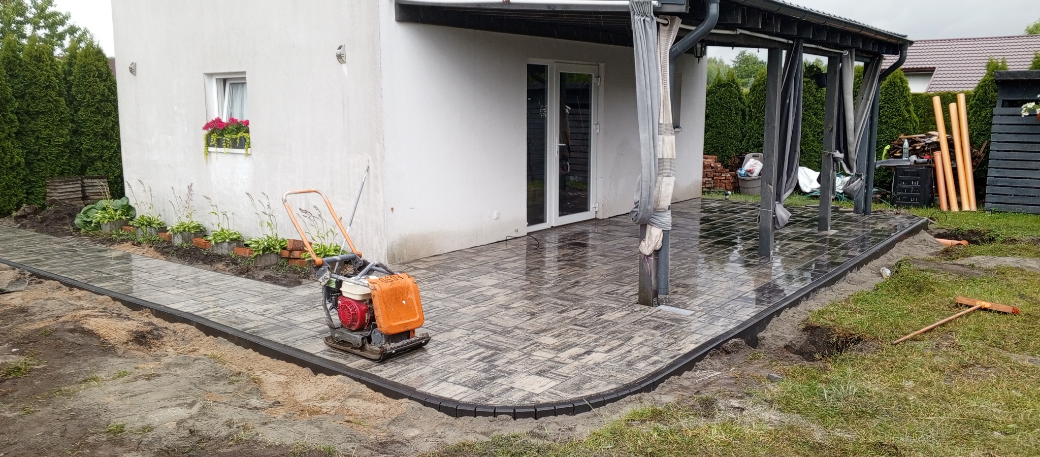 Świeżo ułożona, mokra kostka brukowa w szaro-czarnych odcieniach, obramowana ciemnym obrzeżem. Na kostce stoi pomarańczowa zagęszczarka. Tło: dom z pergolą i zieleń.