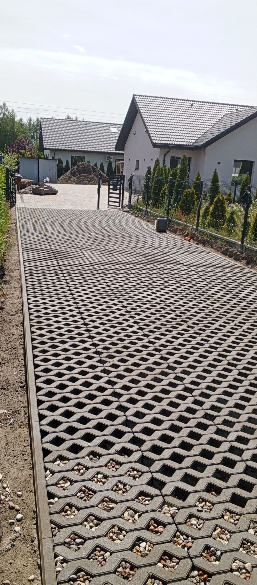 Szara ażurowa nawierzchnia z kamieniami, prowadząca do domu z ciemną dachówką i ogrodzeniem z siatki. Widok na podwórko w słoneczny dzień.
