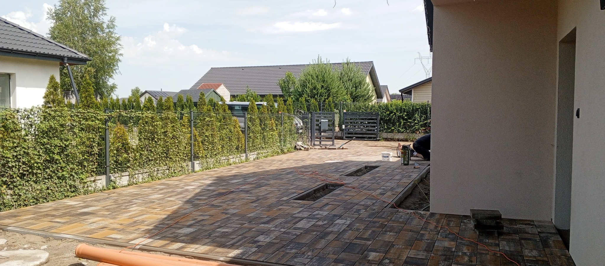 Ułożona kostka brukowa na podjeździe, widoczne elementy montażu, ogrodzenie panelowe z zielenią, domy w tle. Częściowo ukończony projekt.