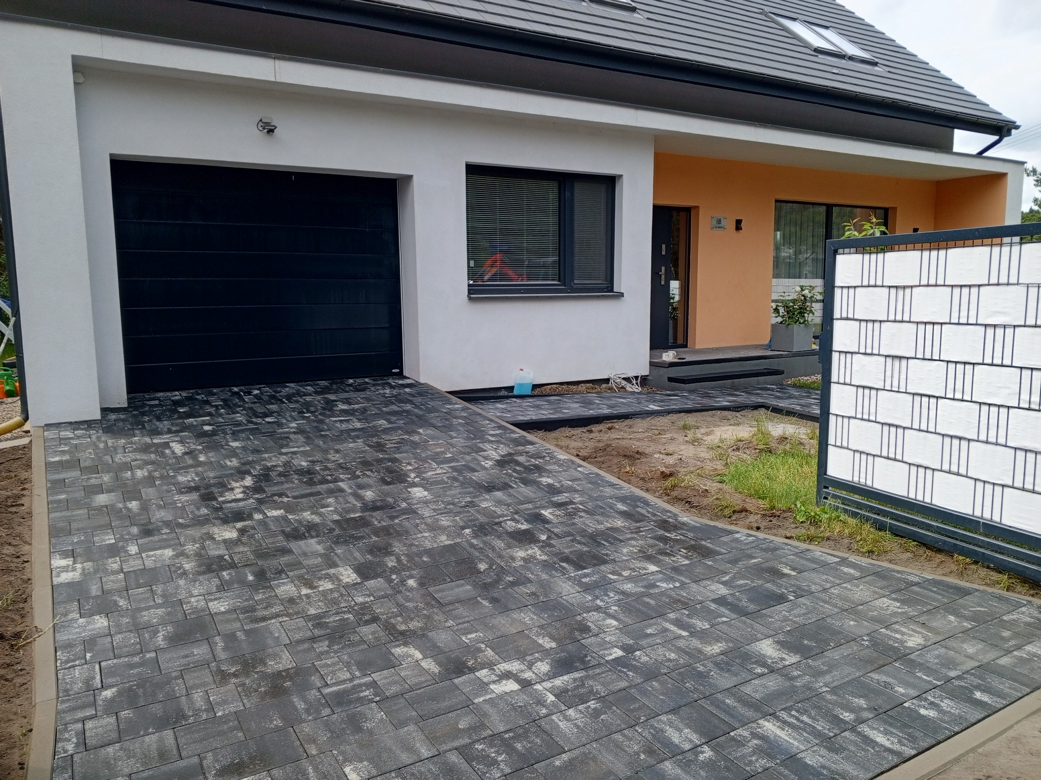 Ułożona kostka brukowa prowadząca do garażu i wejścia domu. Szara kostka o nieregularnym wzorze. Widoczny fragment ogrodzenia panelowego i elewacja domu z czarną bramą garażową.