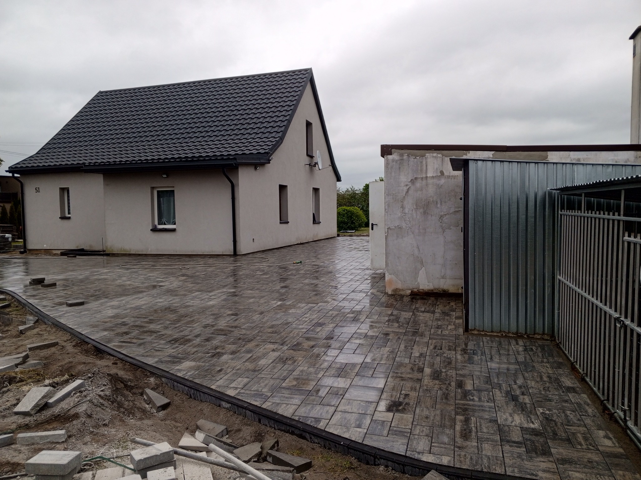 Ułożona kostka brukowa na podjeździe domu z ciemnym dachem, widoczne niedokończone obrzeża i stos kostki. Pochmurne niebo w tle.