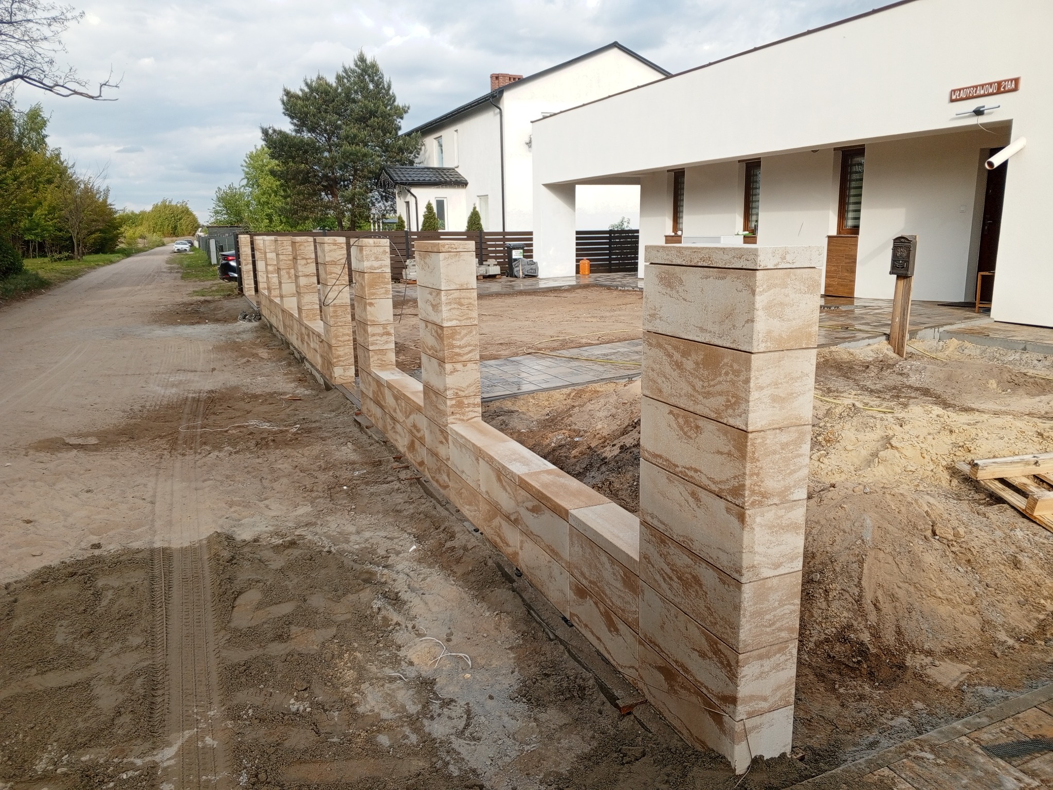 Wznoszenie ogrodzenia z bloków piaskowca na posesji. Widoczny fragment konstrukcji w trakcie budowy, z elementami domu w tle. Surowy teren wokół.