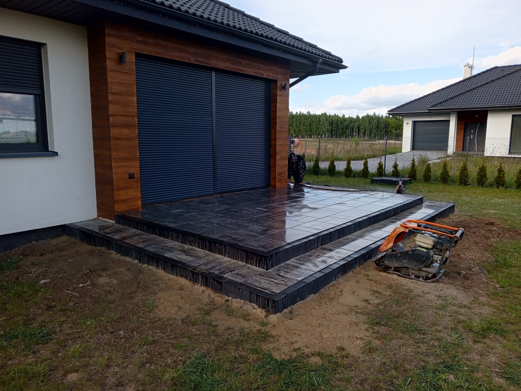 Nowoczesny taras z ciemnej kostki brukowej przy budynku z drewnianą elewacją. Widoczna ubijarka do kostki brukowej. W tle dom i zieleń.