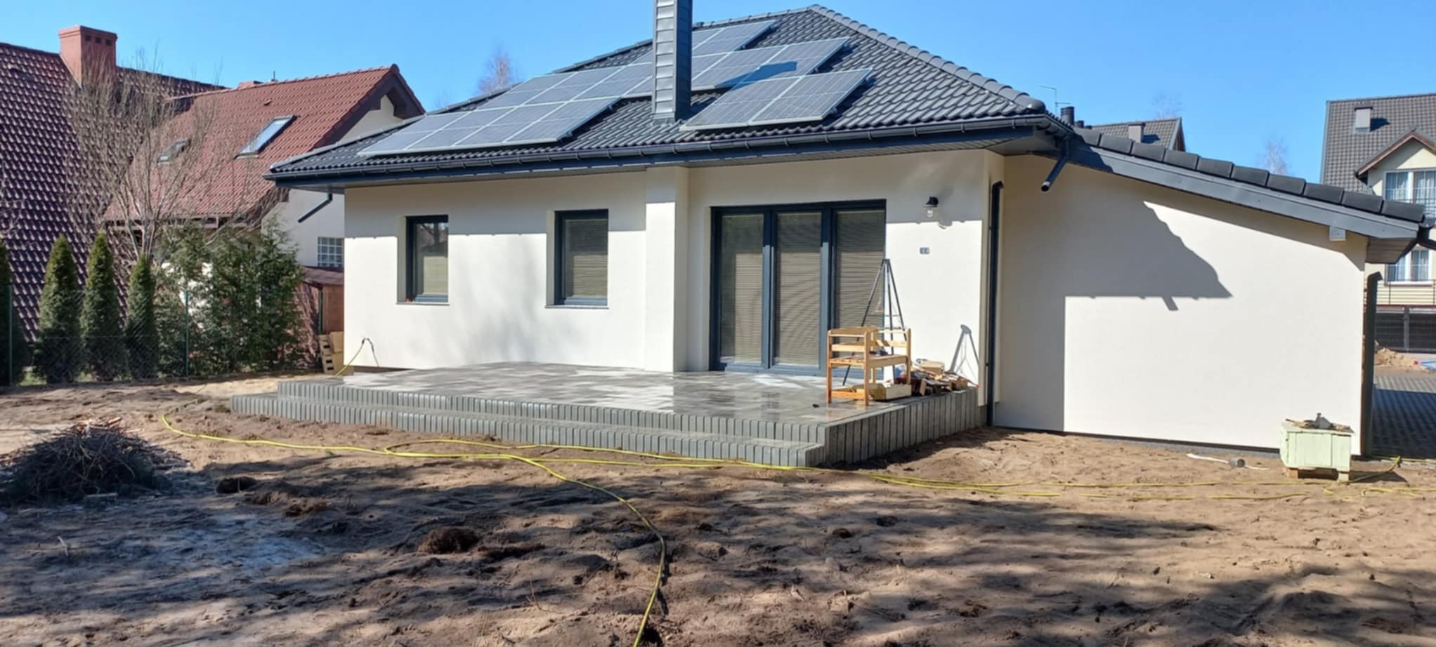 Nowoczesny, parterowy dom z szarym dachem i panelami słonecznymi. Taras z kostki brukowej z szarymi schodami. Wokół widoczna ziemia przygotowana pod zagospodarowanie.