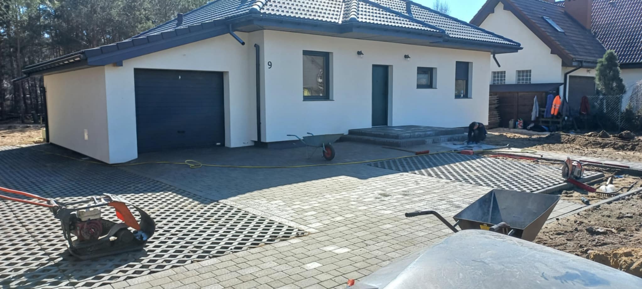 Układanie ażurowej kostki brukowej na podjeździe domu jednorodzinnego. Widoczna ubijarka, taczki i pracownik w trakcie wykonywania prac.