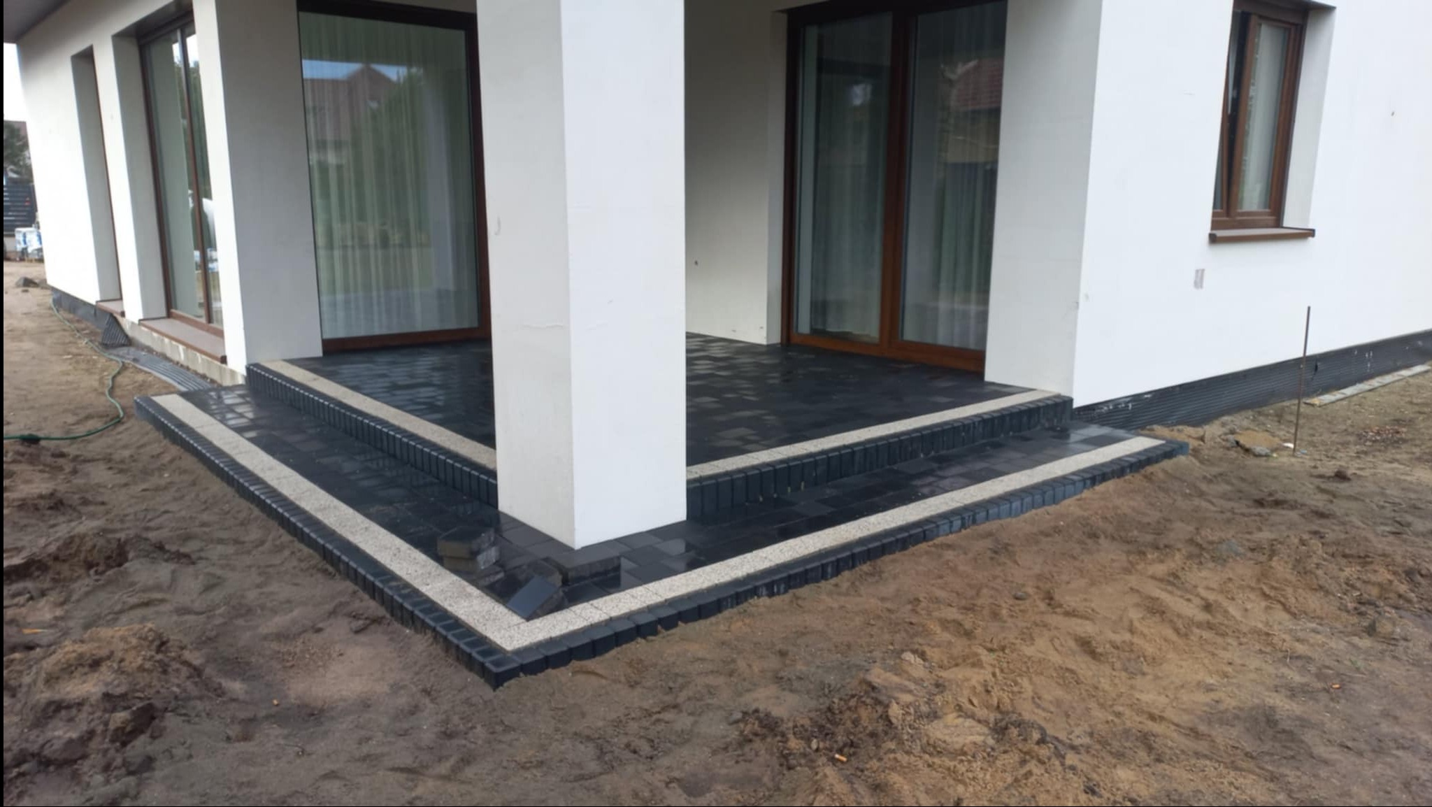 Nowoczesne schody wejściowe z ciemnej kostki brukowej z jasną obwódką, prowadzące na taras domu z białymi kolumnami. Widoczne niedokończone prace ziemne.
