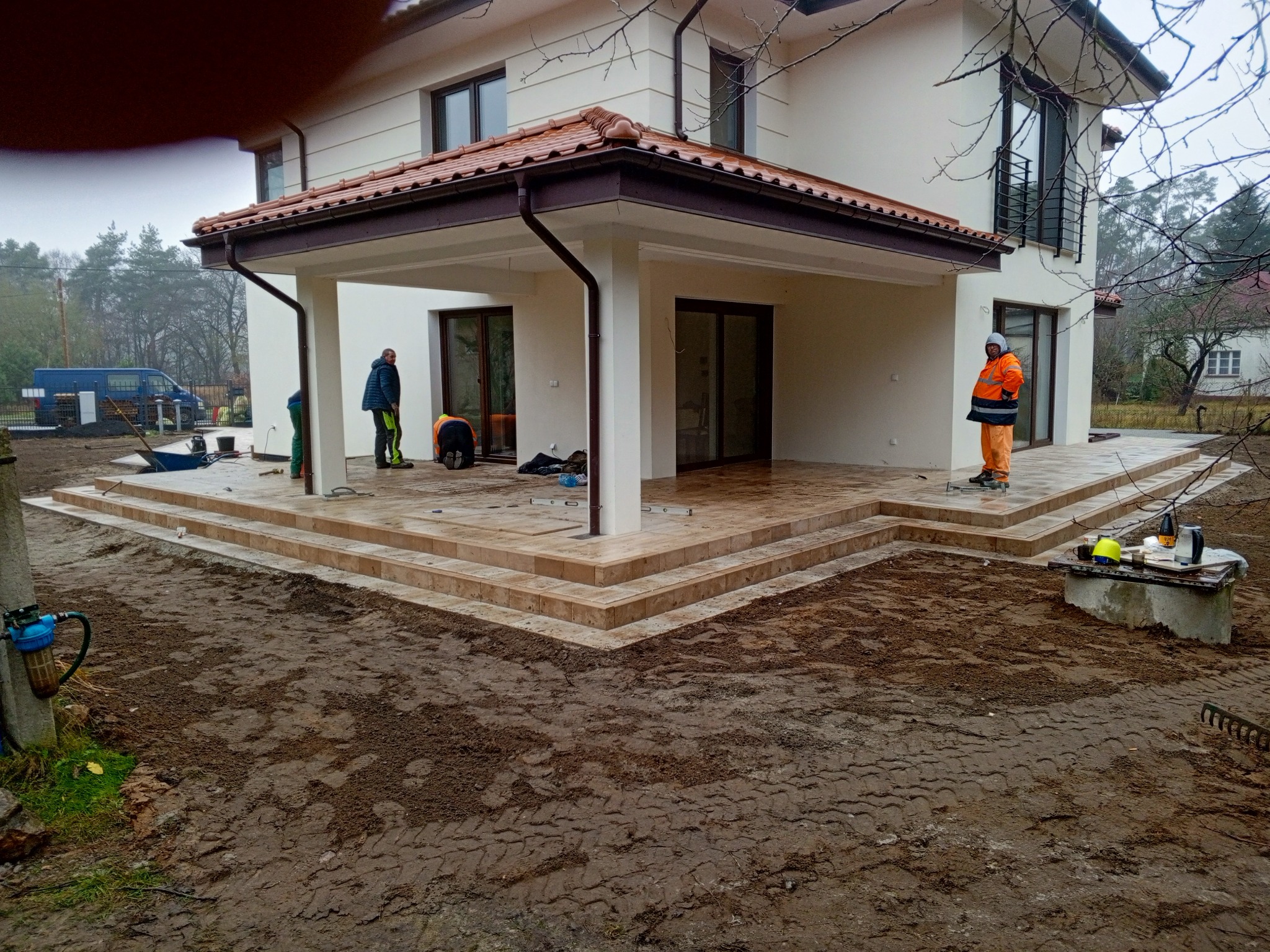 Układanie kostki brukowej na tarasie domu jednorodzinnego. Widoczni pracownicy w trakcie wykonywania prac, kostka w odcieniach beżu, brązowe elementy wykończenia.