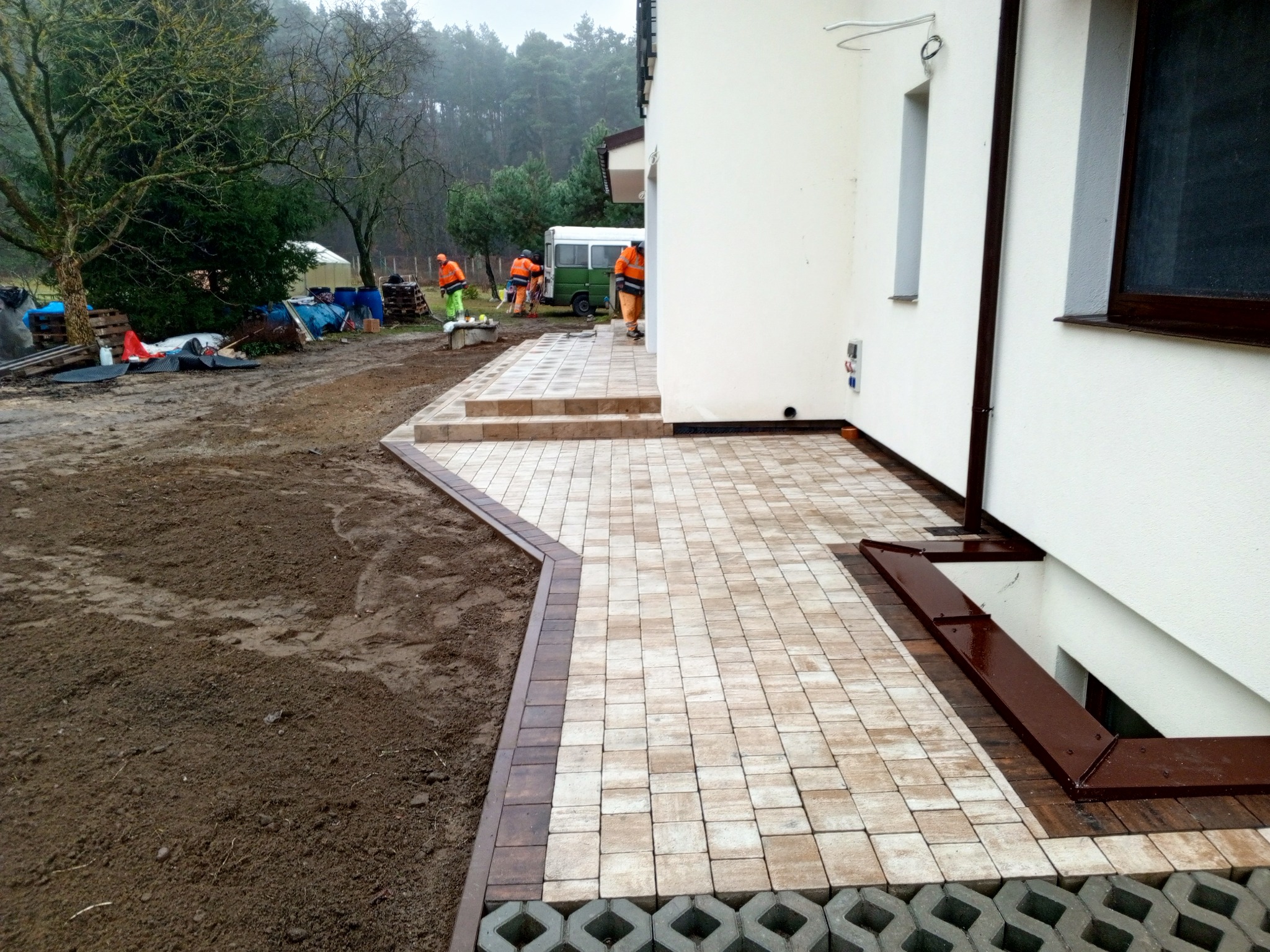 Jasna kostka brukowa ułożona przy białym budynku, z ciemnobrązowym obramowaniem. W tle pracownicy w pomarańczowych strojach i zielony bus.