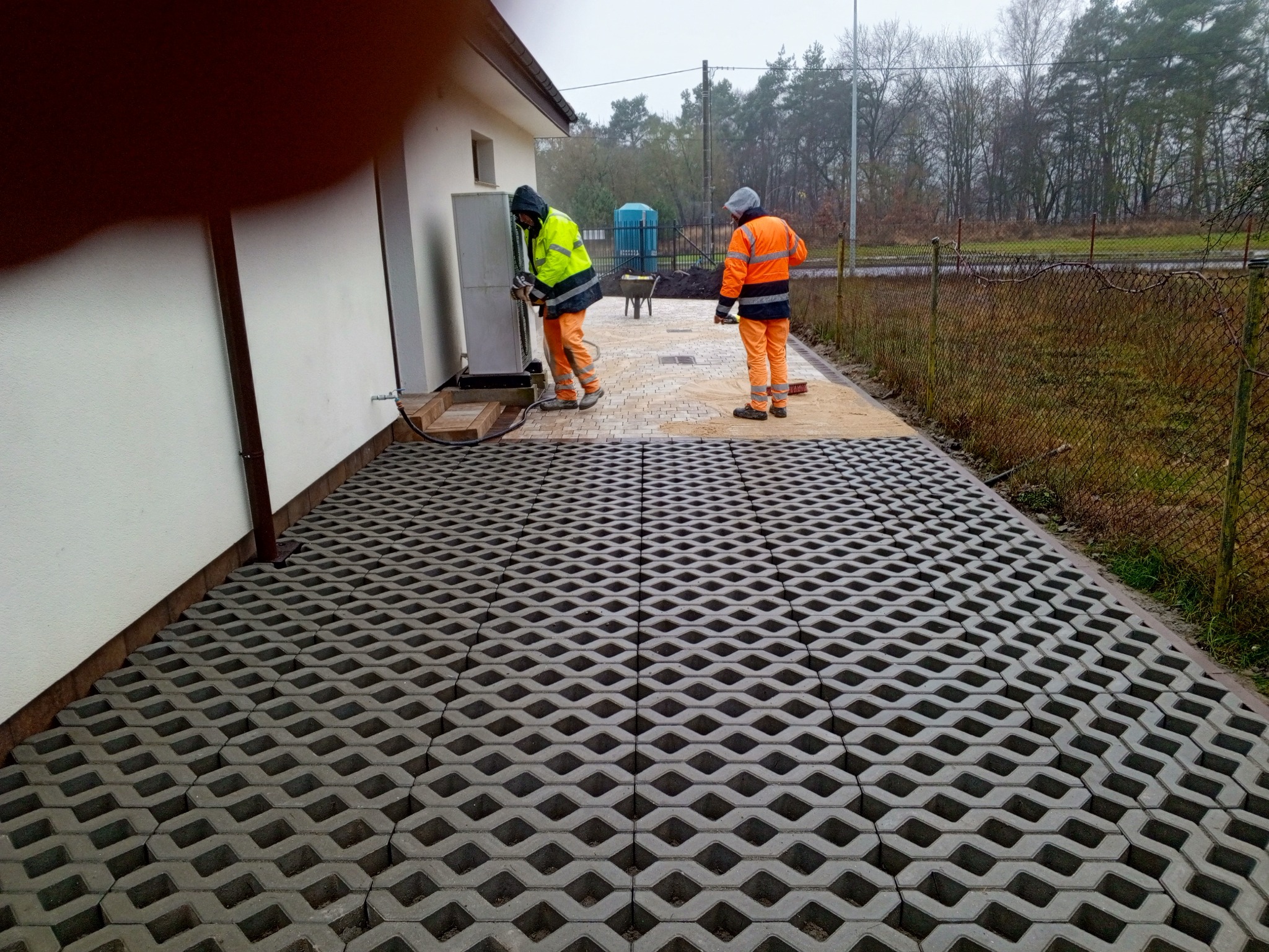 Ułożona szara ażurowa kostka brukowa przy budynku. Dwóch pracowników w odblaskowych ubraniach montuje urządzenie na zewnątrz. W tle widoczne drzewa i ogrodzenie.