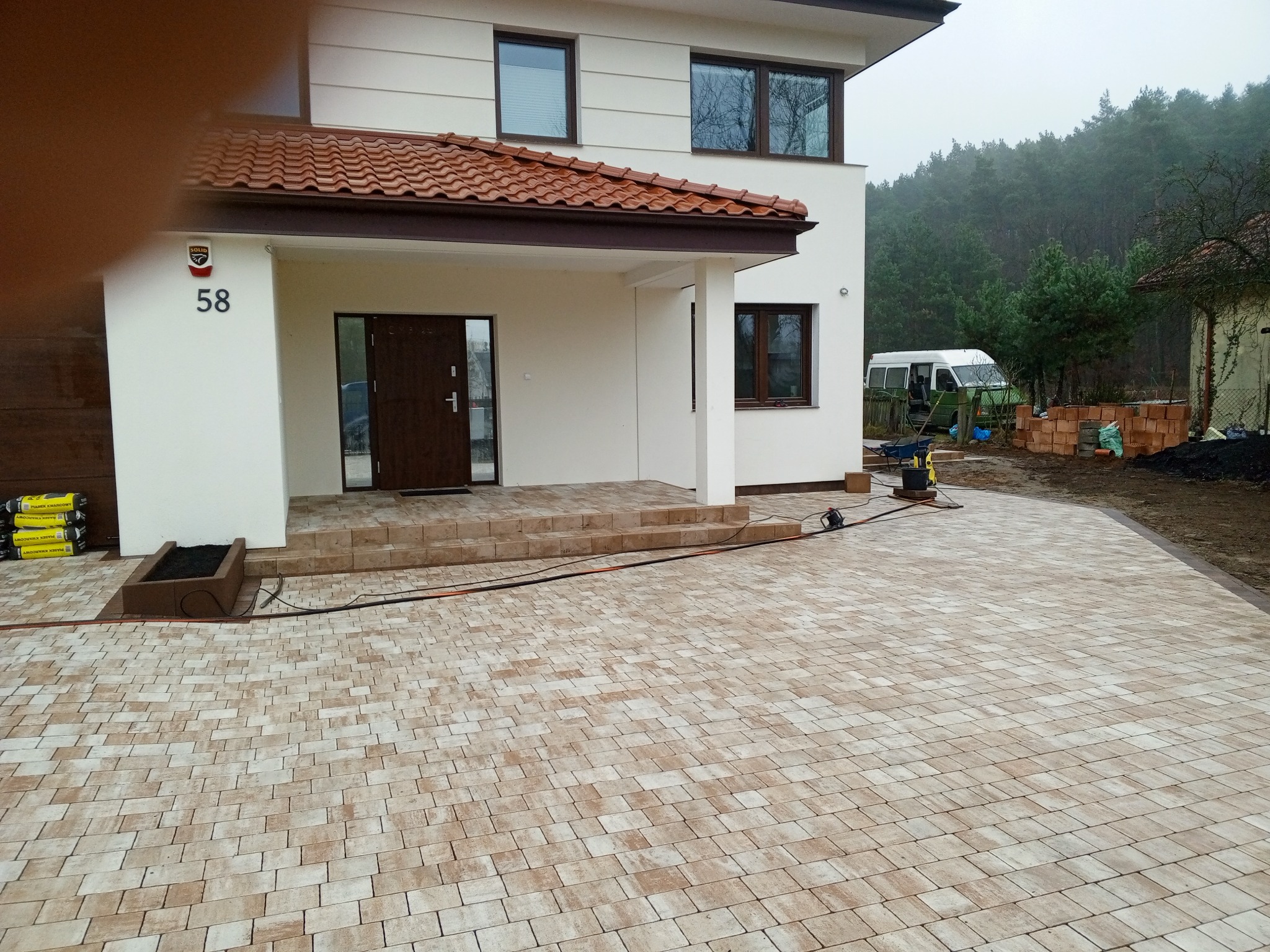 Ułożona kostka brukowa przed domem z beżowo-brązowej kostki. Widoczny fragment budynku z numerem 58 i fragment lasu w tle. Po prawej stronie leżą cegły.