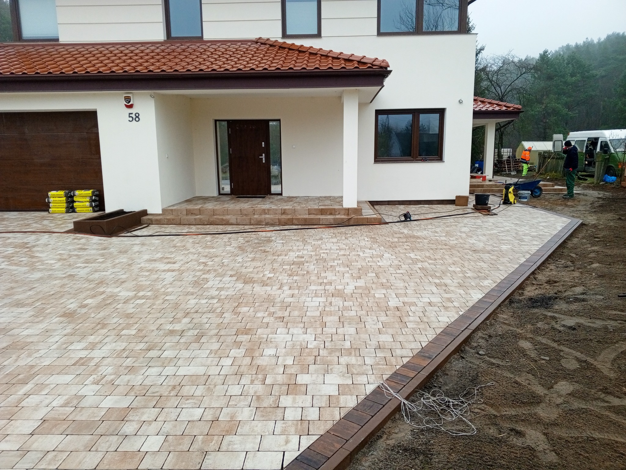 Nowoczesny podjazd z kostki brukowej w odcieniach beżu i brązu, prowadzący do domu z brązowymi akcentami. Widoczne narzędzia i pracownicy w tle.