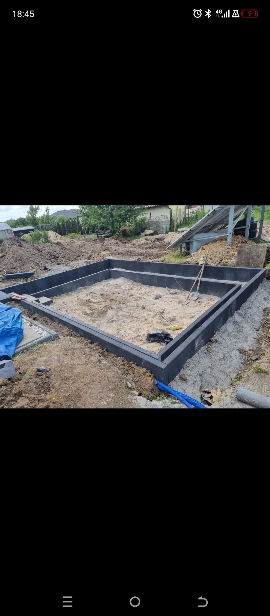 Fundament pod budynek w trakcie budowy, wypełniony piaskiem, z widocznymi narzędziami i elementami instalacji. Otoczenie: trawa, drzewa, domy.
