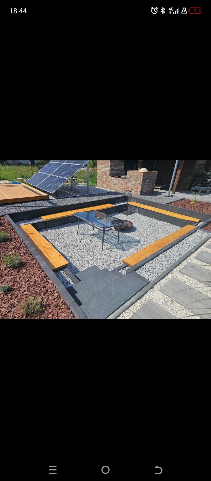 Nowoczesna aranżacja ogrodu z paleniskiem otoczonym kamieniami, drewnianymi ławkami i kostką brukową. W tle widoczny panel słoneczny i fragment budynku z cegły.