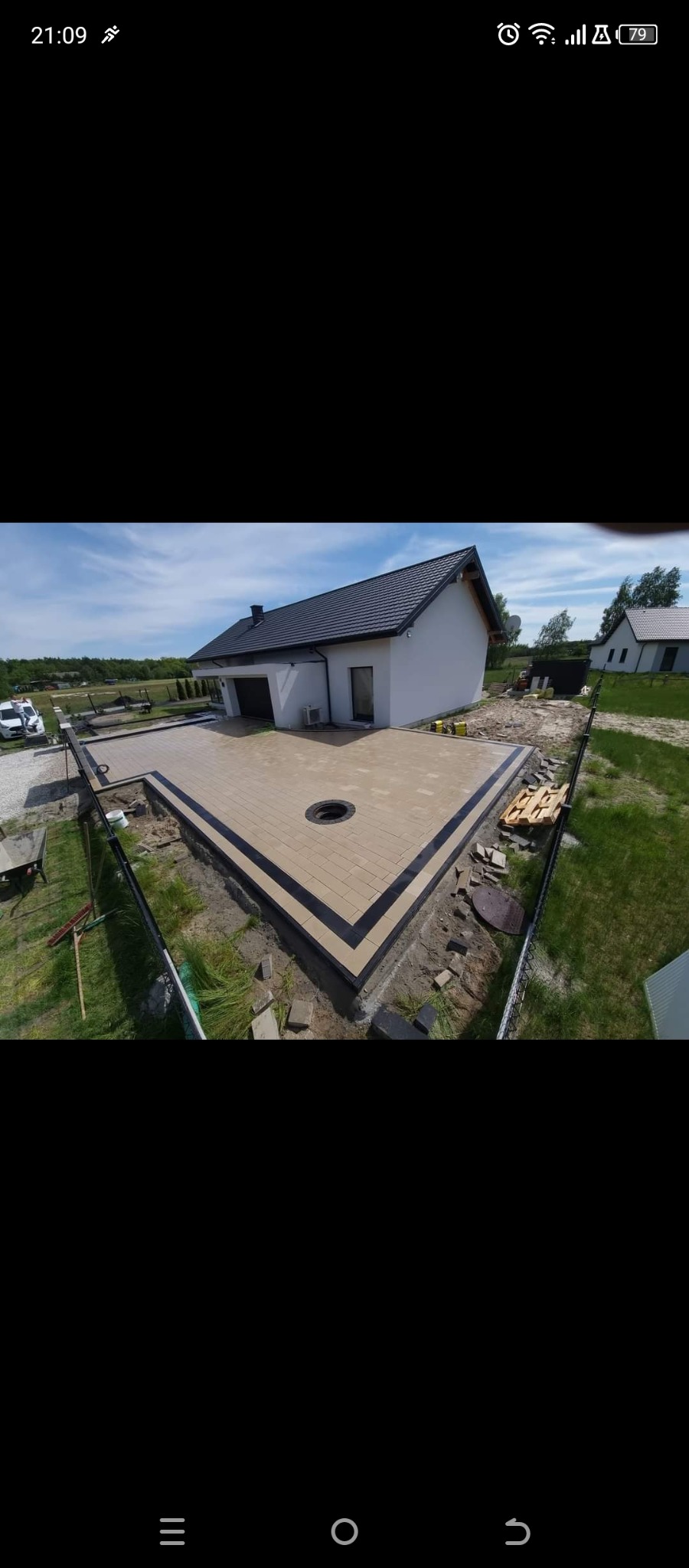 Nowoczesny taras z jasnej kostki brukowej obramowany ciemnym pasem, z okrągłym otworem, otoczony zielenią i domem w tle. Widok z góry.