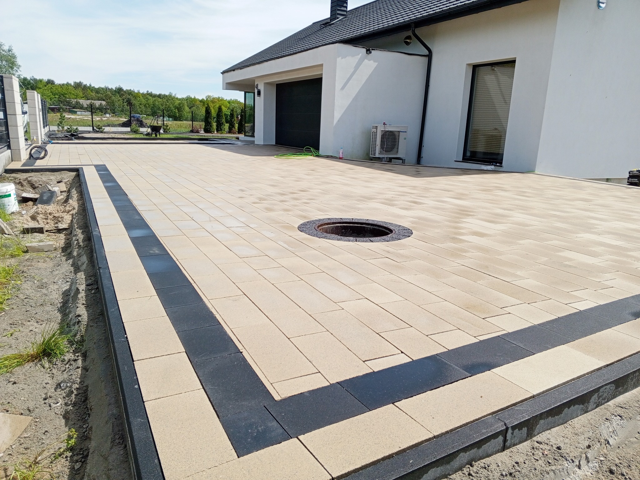 Jasna kostka brukowa z ciemną obwódką i studzienką na podjeździe do nowoczesnego domu z garażem, w słoneczny dzień.