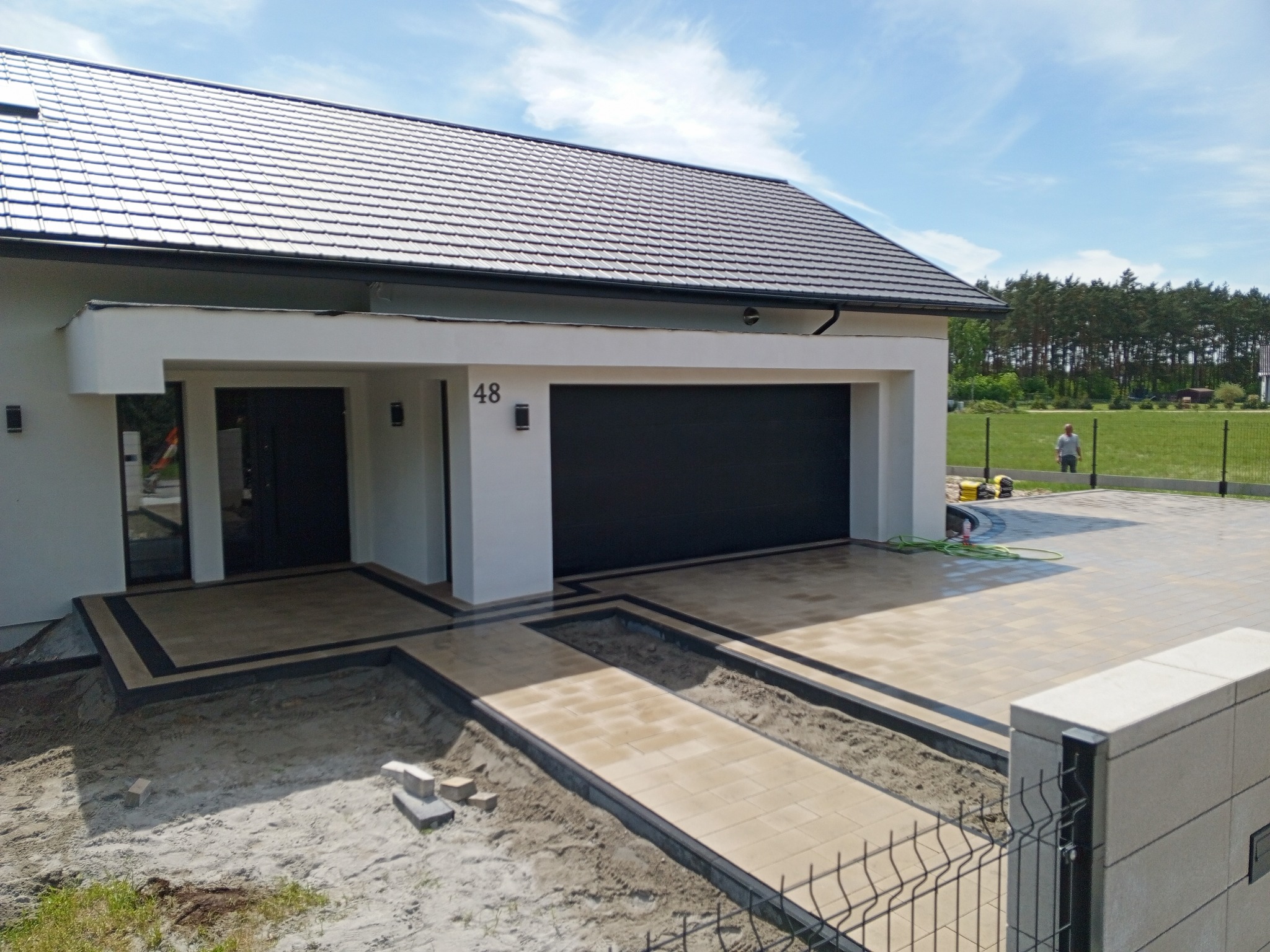 Nowoczesny podjazd z kostki brukowej w jasnych odcieniach, obrzeża w kolorze antracyt. Widoczny fragment domu z numerem 48 i brama garażowa. Prace w toku.
