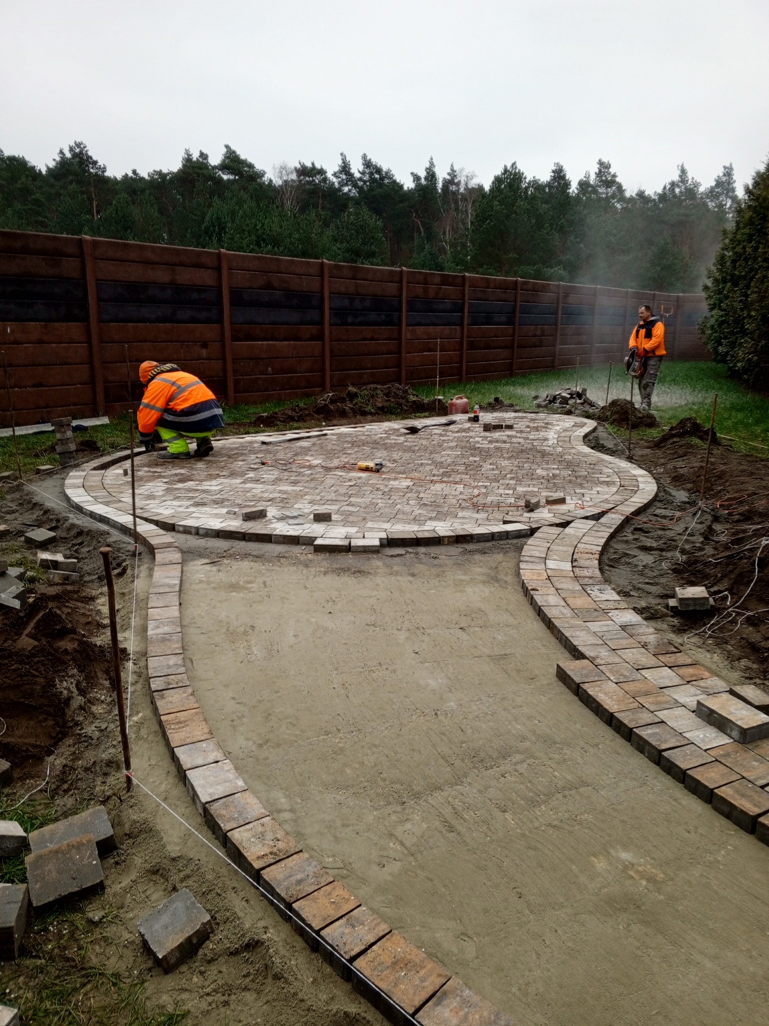 Prace brukarskie w toku: układanie kostki na ścieżce i okrągłym patio. Dwóch pracowników w odblaskowych ubraniach, drewniany płot w tle. Widoczne narzędzia i materiały.