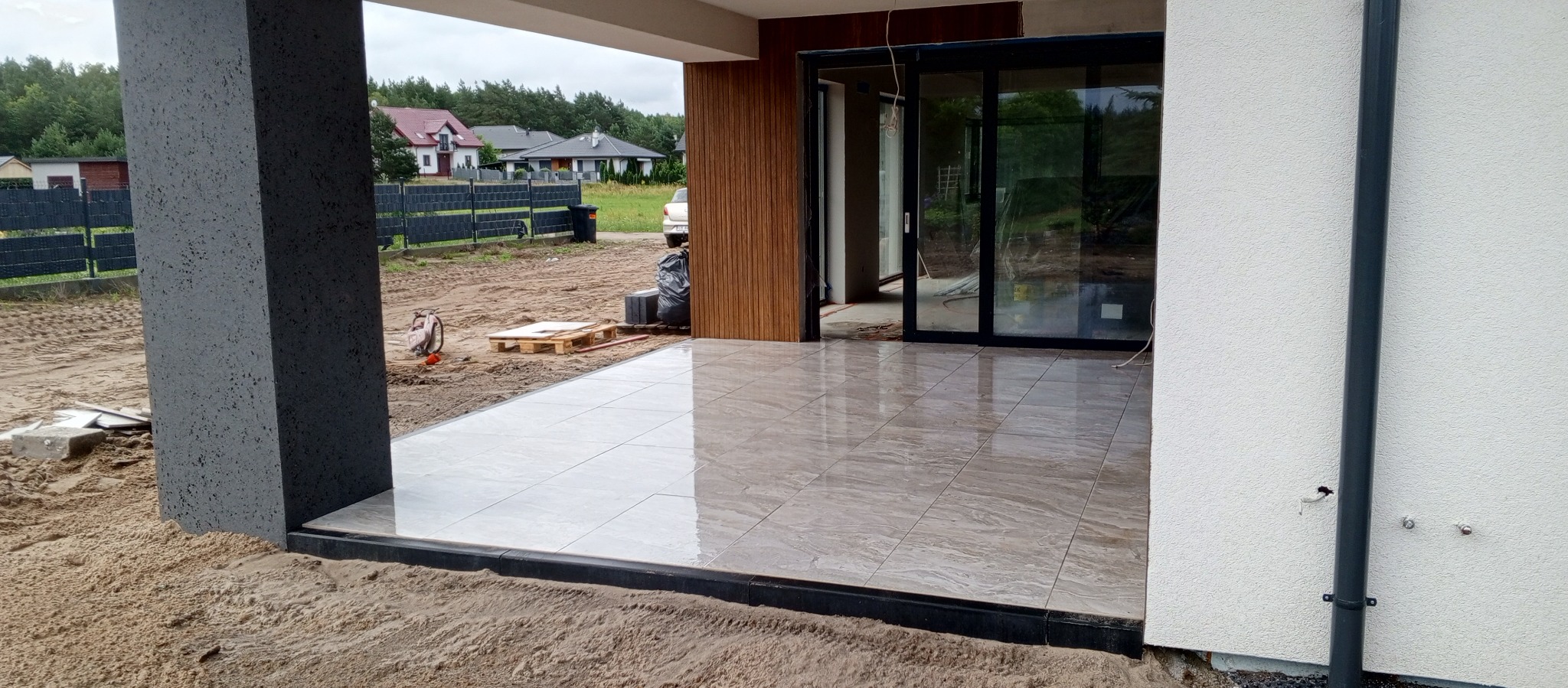 Jasne płytki tarasowe z ciemną obwódką, świeżo ułożone na zewnątrz budynku. Widoczne narzędzia i elementy wykończeniowe. W tle domy jednorodzinne.