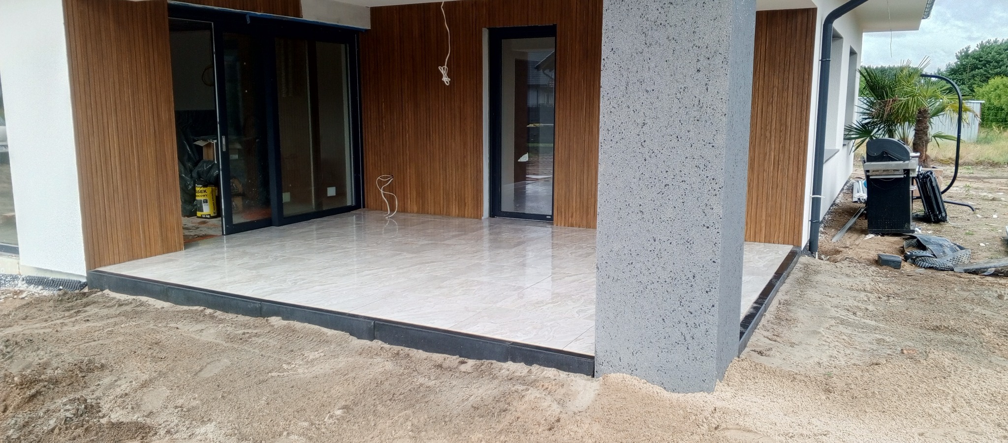 Jasne płytki na tarasie z obrzeżem, widok na kolumnę, okna i przesuwne drzwi. W tle grill i palma. Piasek wokół tarasu wskazuje na prace wykończeniowe.