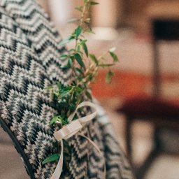 Studio Salach - Krzesło z szaro-białym wzorem, ozdobione gałązką zieleni i kremową wstążką, prawdopodobnie dekoracja ślubna. Tło rozmyte, w ciepłych tonach.