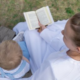Studio Salach - Dziewczynka w białej albie i wianku czyta książkę, siedząc na drewnianych deskach obok małego dziecka w błękitnych ubraniach. Widok z góry na uroczystość religijną.
