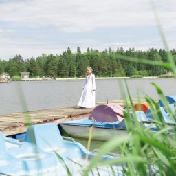 Studio Salach - Dziewczyna w białej sukni stoi na drewnianym pomoście nad jeziorem, obok kolorowych rowerów wodnych. W tle las i błękitne niebo z chmurami.