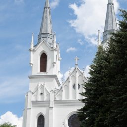 Studio Salach - Biały kościół z dwiema wieżami zwieńczonymi krzyżami, częściowo zasłonięty przez zielone drzewo. Błękitne niebo z chmurami w tle.