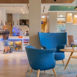 Studio Salach - Wnętrze hotelowe z recepcją w tle i strefą wypoczynkową z niebieskimi fotelami i drewnianymi stolikami. Widoczne przeszklone drzwi i elementy dekoracyjne.