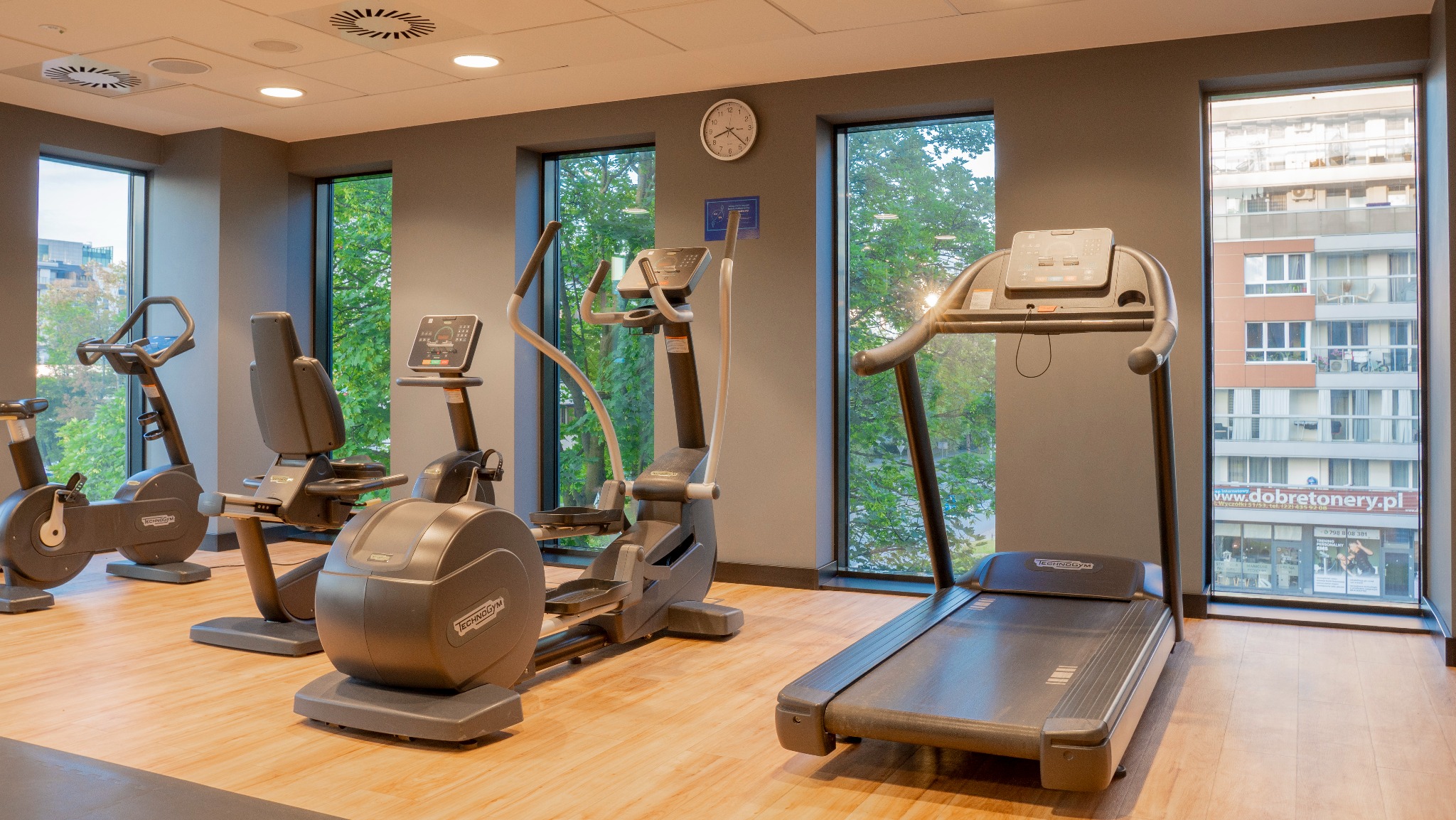 Jasne wnętrze siłowni z nowoczesnym sprzętem Technogym: bieżnia, rowery stacjonarne i orbitrek, z widokiem na zieleń i budynki przez duże okna.