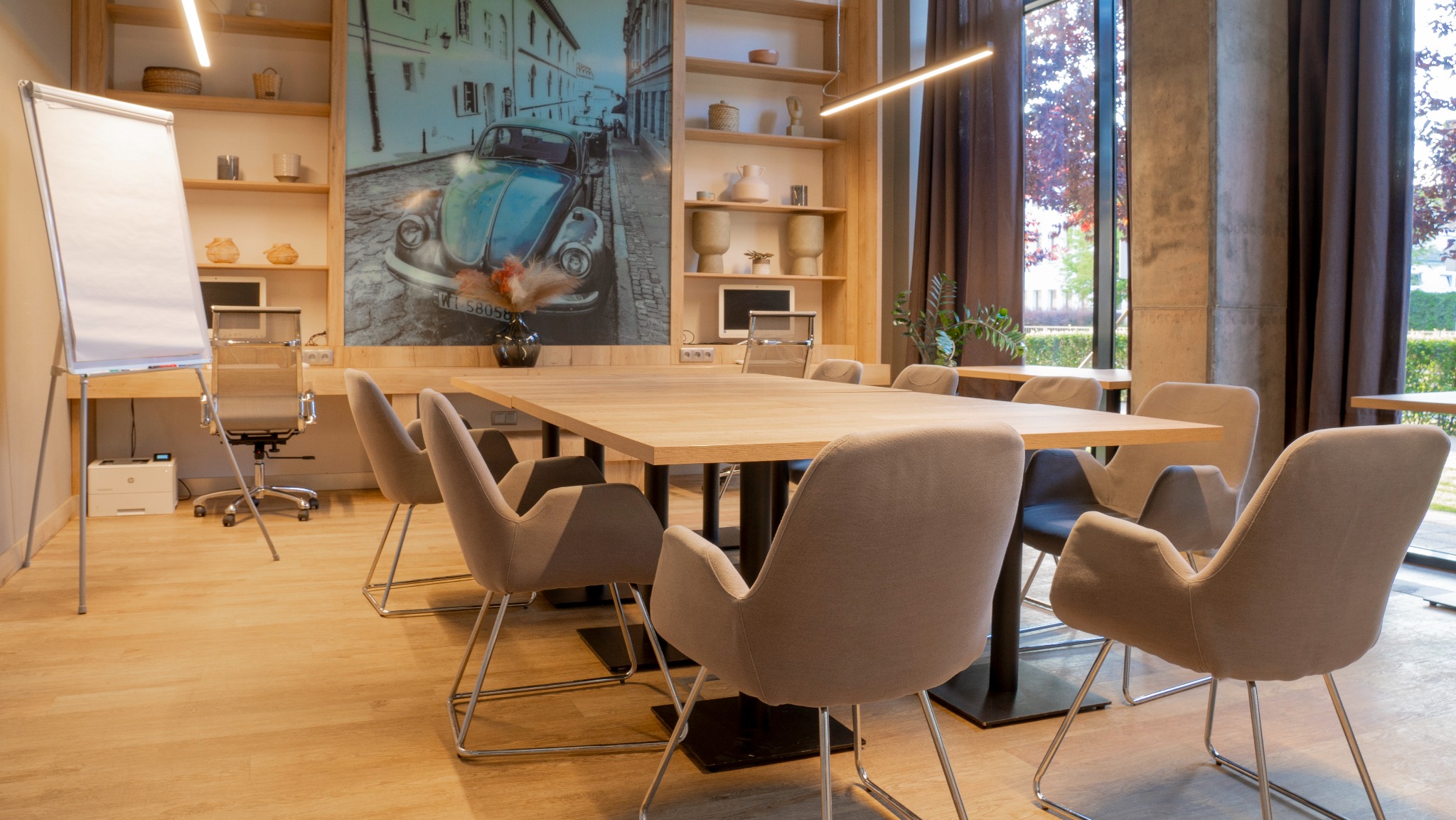 Jasne biuro z drewnianym stołem konferencyjnym i krzesłami. W tle regał z dekoracjami i obraz z motywem auta. Duże okno z widokiem na zieleń.