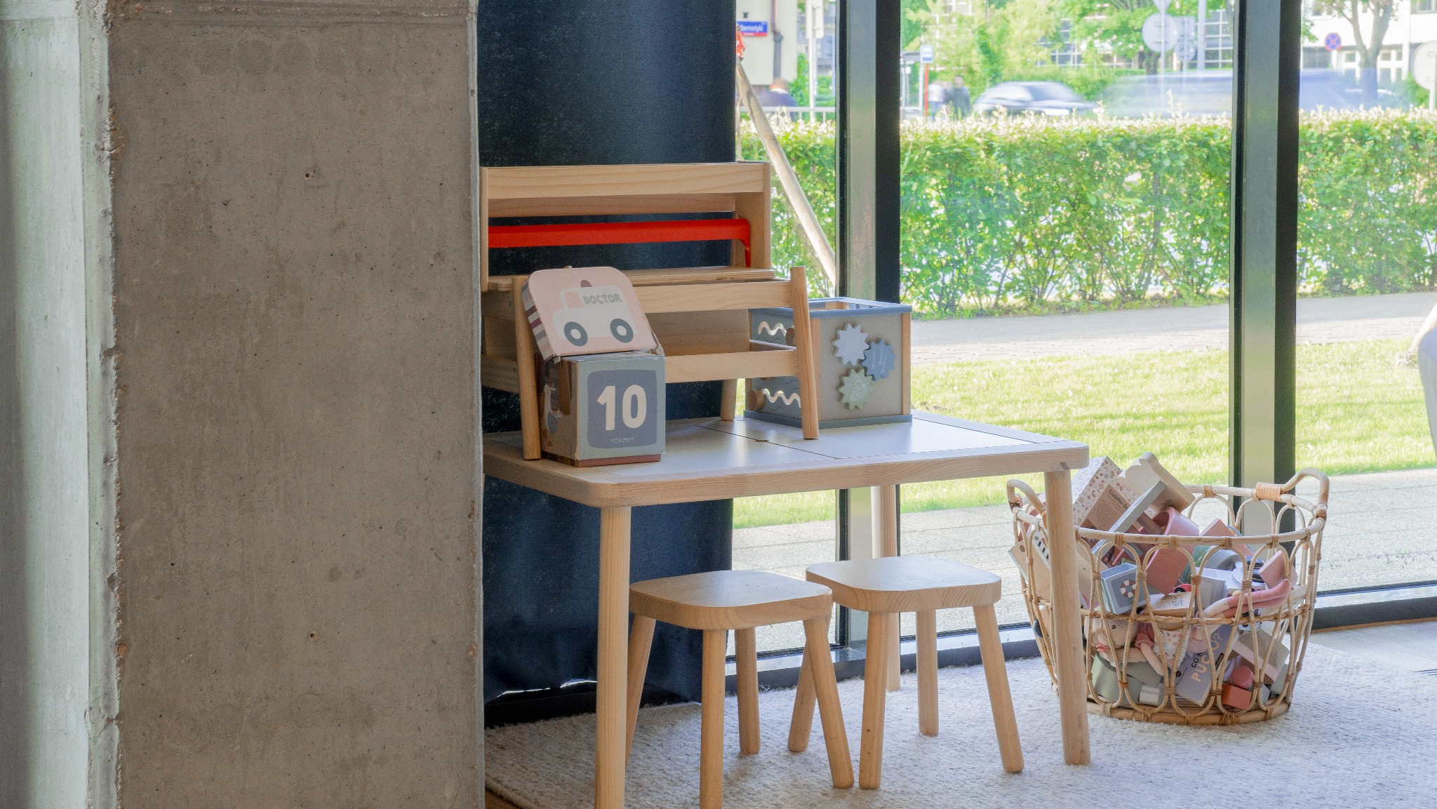 Jasny, drewniany stolik dziecięcy z krzesełkami i zabawkami na tle okna z widokiem na zieleń. W koszu obok stolika dodatkowe zabawki.