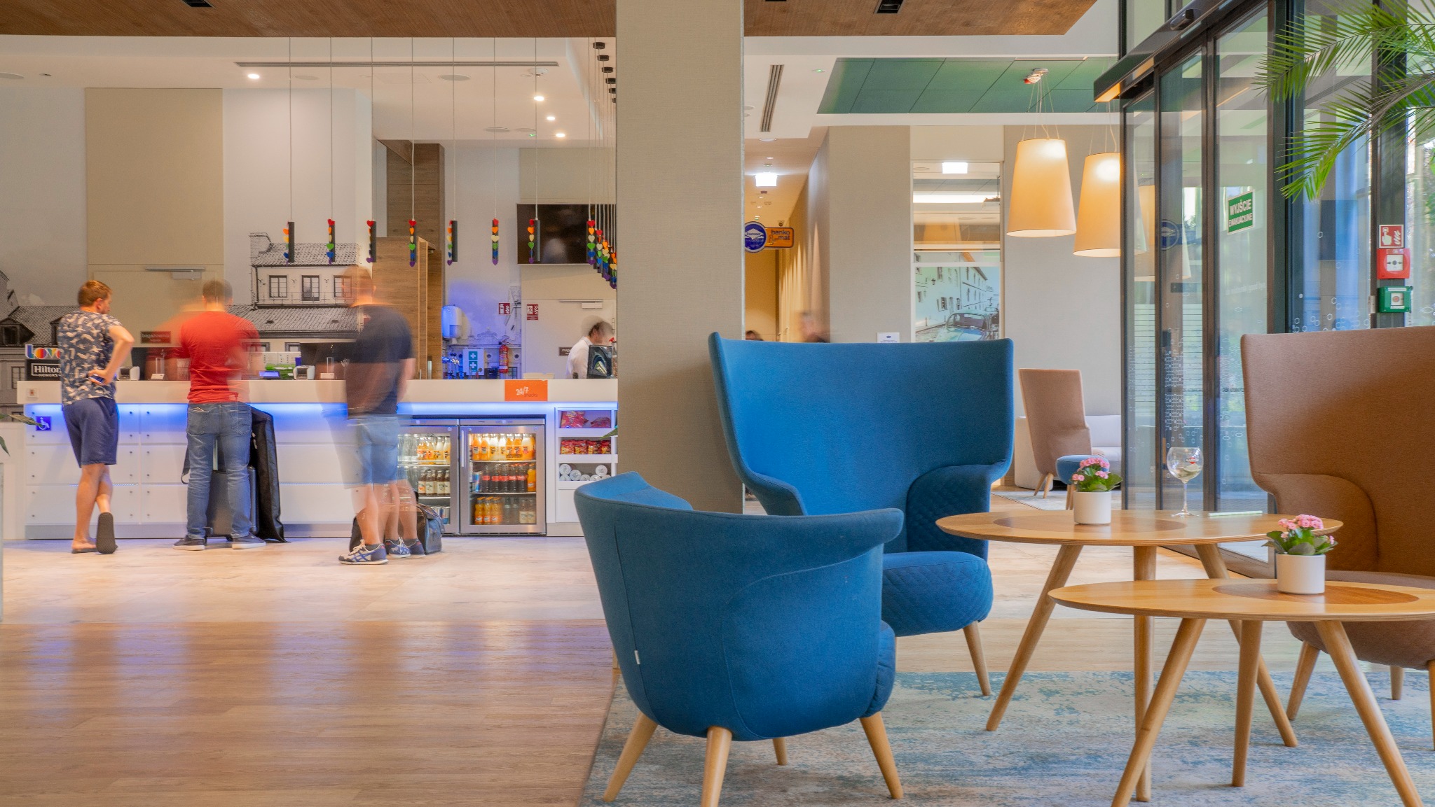 Wnętrze hotelowe z recepcją w tle i strefą wypoczynkową z niebieskimi fotelami i drewnianymi stolikami. Widoczne przeszklone drzwi i elementy dekoracyjne.