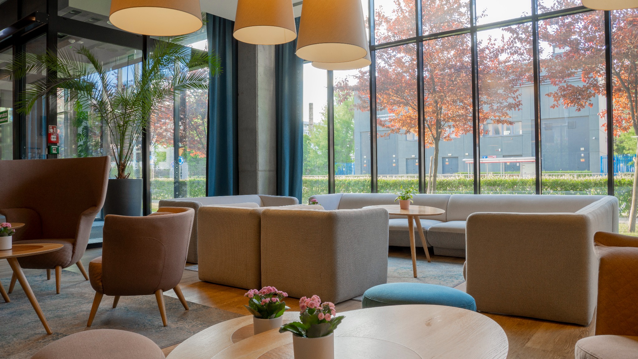 Jasne, przestronne wnętrze poczekalni z wygodnymi fotelami, sofami i stolikami kawowymi. Duże okna z widokiem na drzewa o czerwonych liściach w otoczeniu budynków.