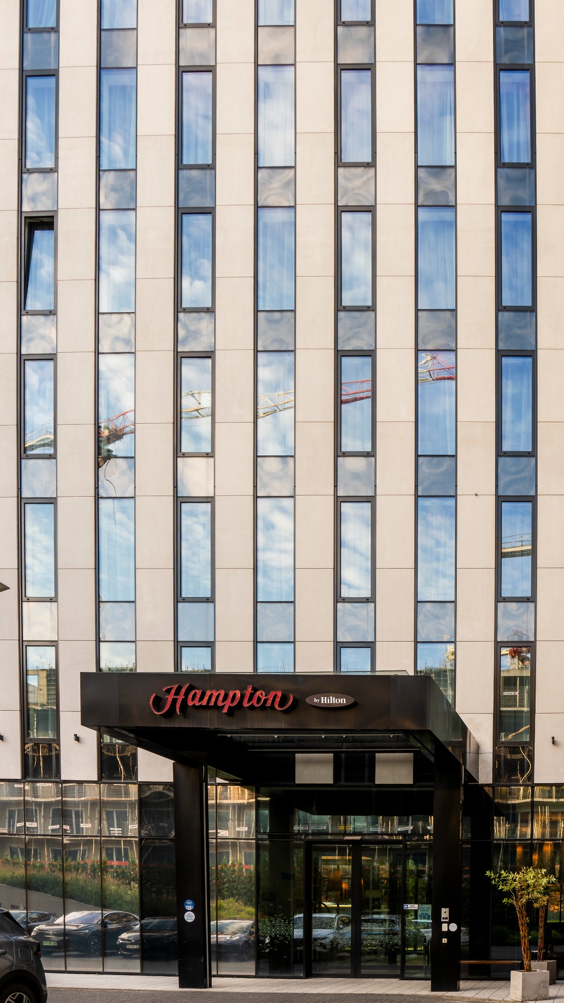 Fasada hotelu Hampton by Hilton z licznymi oknami odbijającymi niebo i dźwig budowlany, widoczny nad wejściem z czarnym baldachimem i czerwonym napisem.