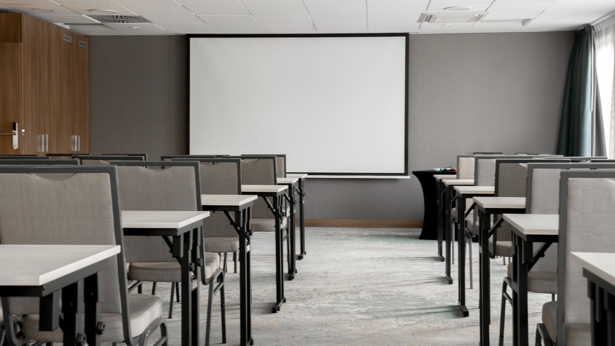 Sala konferencyjna z rzędami krzeseł i stołów, ekran projekcyjny na ścianie, nowoczesny wystrój wnętrza, stonowana kolorystyka, oświetlenie sufitowe.