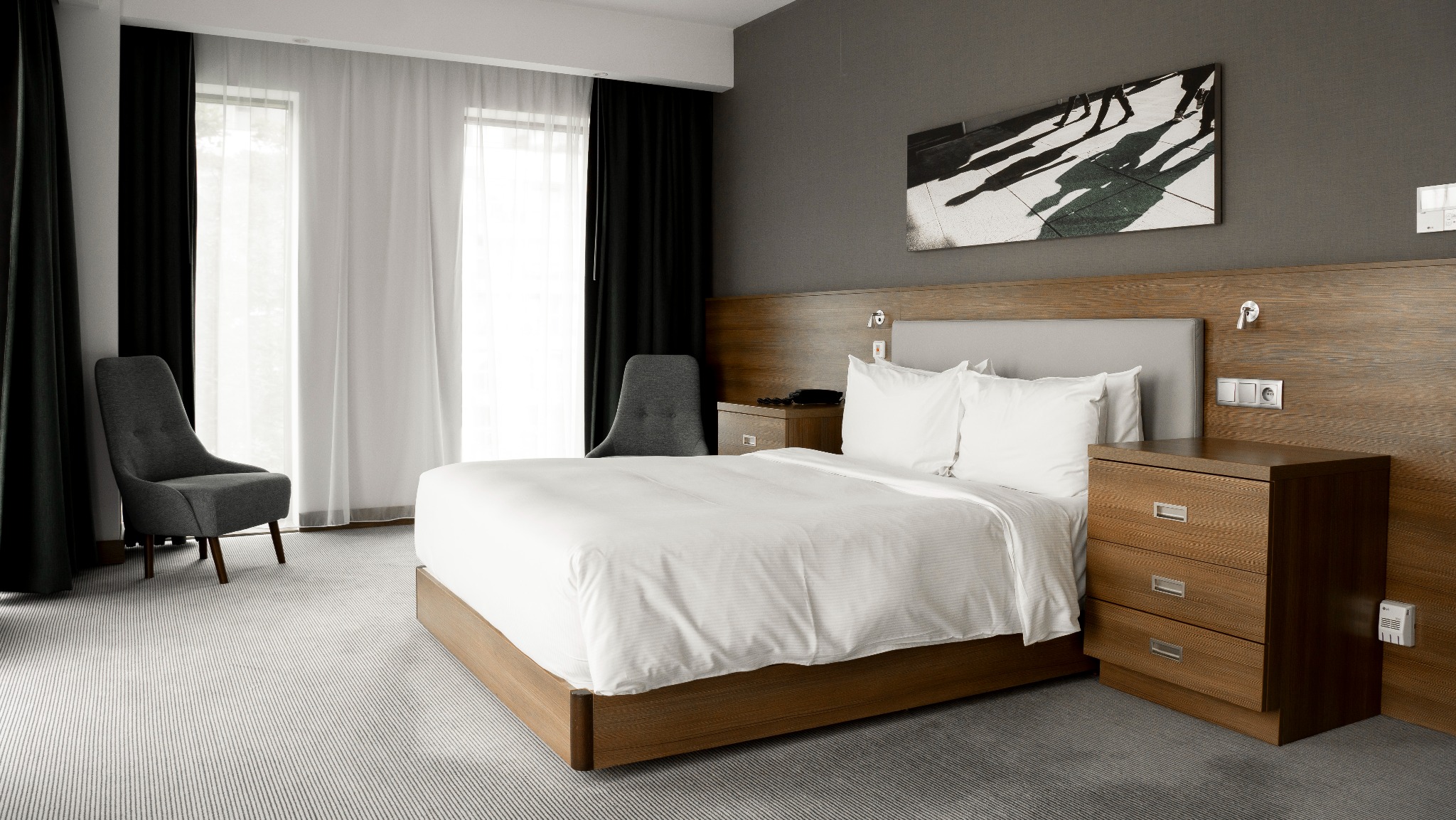 Nowoczesna sypialnia hotelowa z dużym łóżkiem, drewnianym wezgłowiem i stolikami nocnymi. Szare ściany, biała pościel i krzesło w rogu. Na ścianie grafika.