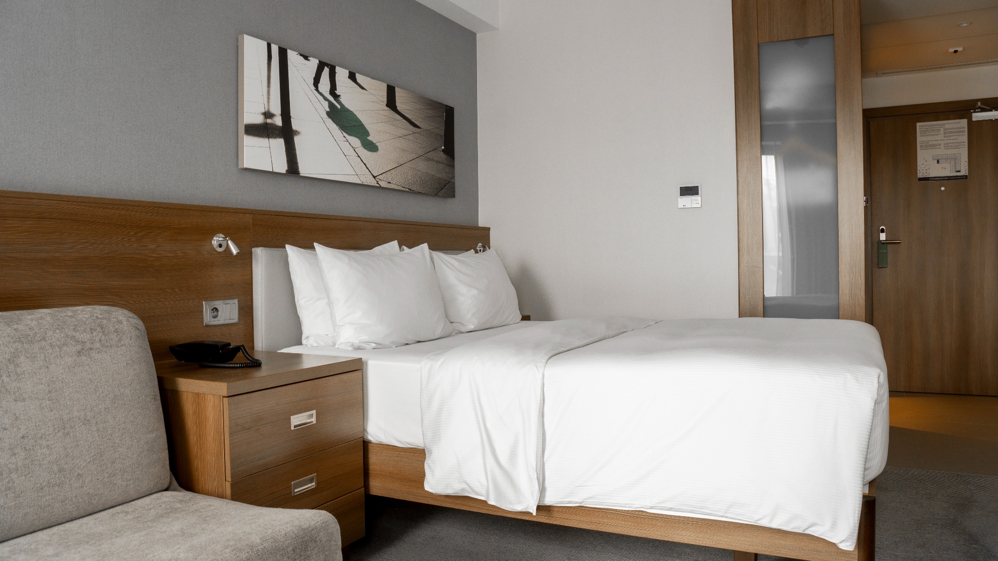 Jasne wnętrze pokoju hotelowego z łóżkiem, szafką nocną z telefonem i krzesłem. Na ścianie grafika przedstawiająca sylwetki ludzi.