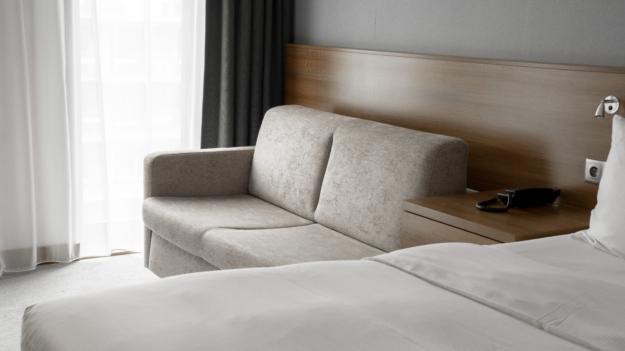 Pokój hotelowy w neutralnych barwach: beżowa sofa z drewnianym zagłówkiem, biała pościel, stolik nocny z telefonem i zasłony w tle. Nowoczesne wnętrze.