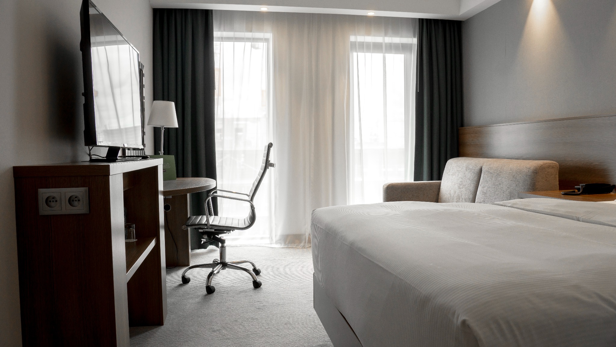 Jasne wnętrze pokoju hotelowego: duże okna z zasłonami, biurko z krzesłem, telewizor na szafce i łóżko z sofą. Minimalistyczny design, stonowana kolorystyka.