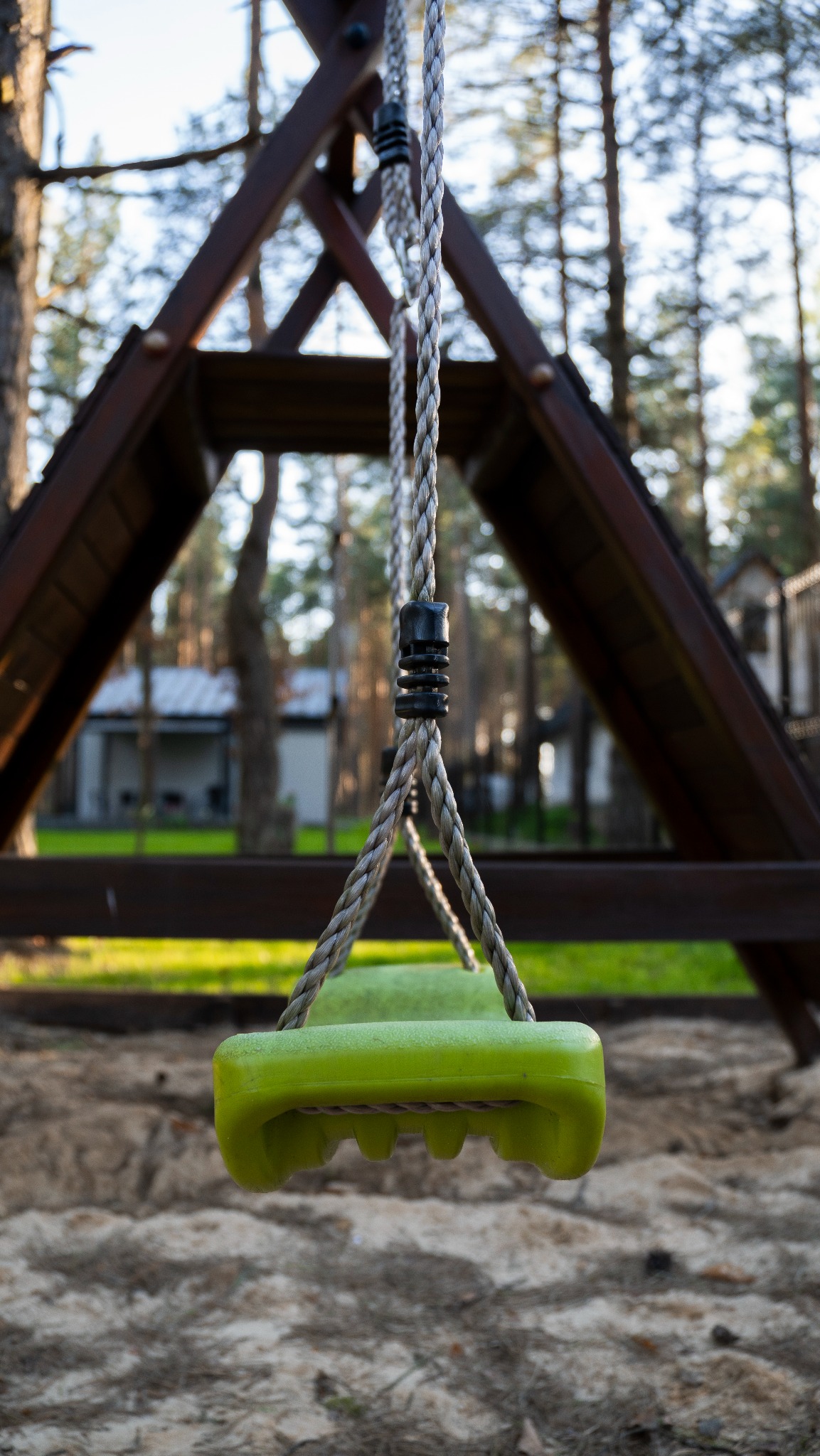 Zielona huśtawka na tle drewnianej konstrukcji i drzew, z widocznymi detalami liny i siedziska, w otoczeniu leśnego krajobrazu i piaszczystego podłoża.