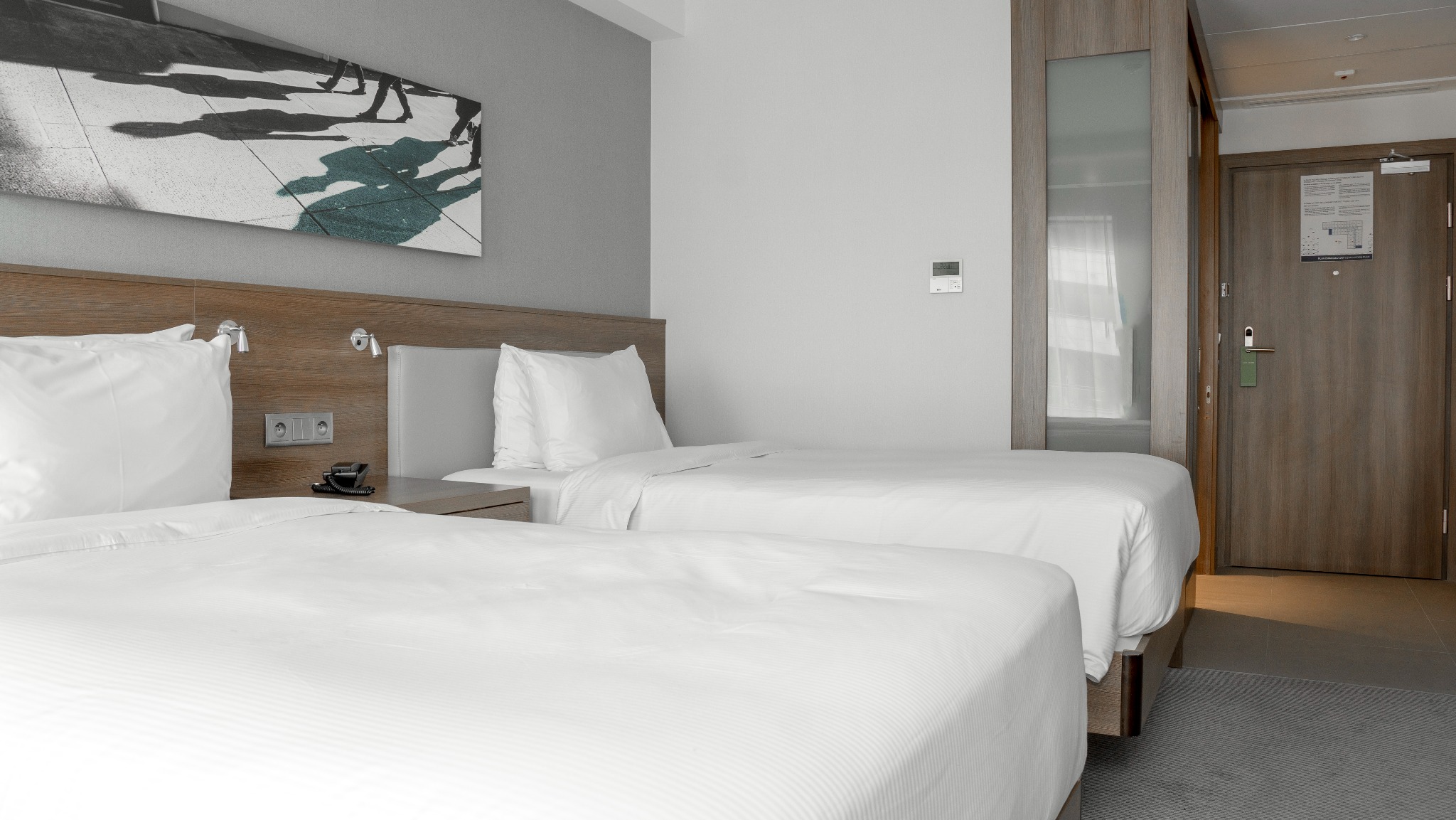 Pokój hotelowy z dwoma pojedynczymi łóżkami, nad którymi wisi nowoczesny obraz. Widoczny stolik nocny z telefonem i gniazdkami.